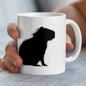 Könnte beinhalten: Weiße Keramik-Tasse mit einer schwarzen Silhouette eines Capybaras. Das Capybara ist in sitzender Position mit detaillierter Fellstruktur dargestellt. Die Tasse hat einen Standardgriff und eine glänzende Oberfläche, geeignet für Heiß- oder Kaltgetränke.