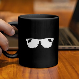 Könnte beinhalten: Eine schwarze Kaffeetasse mit einem weißen Sonnenbrillen-Design. Die Tasse wird von einer Hand gehalten. Im Hintergrund ist ein Laptop auf einer Holzoberfläche zu sehen.