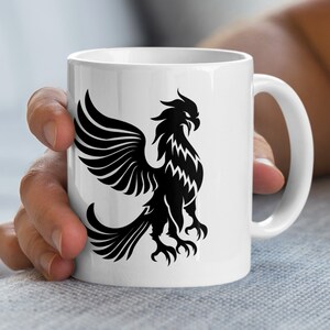 Könnte beinhalten: Weiße Keramiktasse mit einem schwarzen Phönix-Design. Der Phönix wird mit ausgebreiteten Flügeln und detaillierten Federn dargestellt. Die Tasse hat einen gebogenen Griff und eine glänzende Oberfläche.
