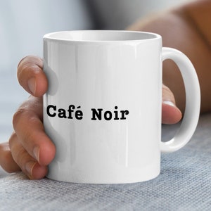 Peut inclure: Tasse à café en céramique blanche avec l'inscription "Café Noir" en noir. La tasse a une anse incurvée et est tenue par une personne. Idéale pour les boissons chaudes.