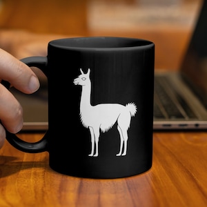 Könnte beinhalten: Schwarze Keramik-Tasse mit einer weißen Lama-Silhouette. Das Lama-Design ist ein einfacher Umriss, der mittig auf der Tasse platziert ist. Die Tasse wird von einer Person gehalten, was auf die Verwendung für Getränke hindeutet.