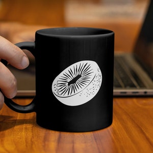 Könnte beinhalten: Schwarze Keramik-Tasse mit einer weißen Illustration einer Kiwi-Fruchtscheibe. Das Design zeigt den Kern und die äußere Textur der Frucht. Die Tasse wird von einer Hand gehalten, mit einem Laptop im Hintergrund.