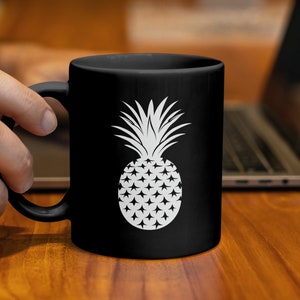 Könnte beinhalten: Schwarze Kaffeetasse mit einem weißen Ananas-Design. Die Ananas-Illustration ist auf der Tasse zentriert und zeigt eine detaillierte Krone und einen strukturierten Körper. Die Tasse wird von einer Person gehalten, im Hintergrund ist ein Laptop zu sehen.
