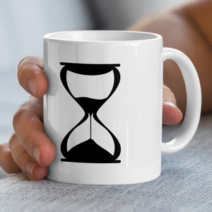 Könnte beinhalten: Weiße Keramik-Tasse mit einem schwarzen Sanduhr-Design. Die Sanduhr zeigt zwei verbundene Kammern, mit Sand, der von oben nach unten fließt. Die Tasse hat einen gebogenen Griff.