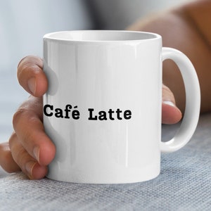 Könnte beinhalten: Weiße Keramik-Kaffeetasse mit dem schwarzen Aufdruck "Café Latte". Die Tasse hat einen gebogenen Henkel und wird von einer Person gehalten.