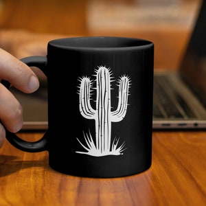 Könnte beinhalten: Schwarze Keramik-Tasse mit einem weißen Kaktus-Design. Die Kaktus-Illustration hat detaillierte Stacheln und eine stilisierte Basis. Die Tasse wird von einer Hand gehalten, im Hintergrund ist ein Laptop zu sehen.