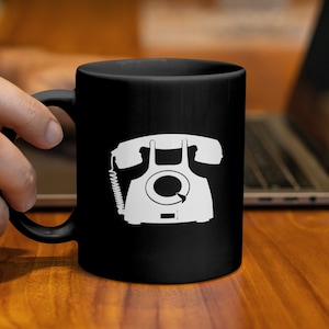 Könnte beinhalten: Schwarze Keramik-Tasse mit einer weißen Grafik eines alten Wählscheibentelefons. Die Tasse hat einen gebogenen Griff und eine zylindrische Form. Das Design ist eine einfache, zweidimensionale Illustration des Telefons.