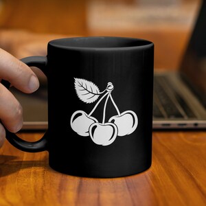 Könnte beinhalten: Schwarze Keramiktasse mit einer weißen Illustration von drei Kirschen und einem Blatt. Die Tasse wird von einer Hand gehalten. Im Hintergrund sind eine Holzoberfläche und ein Laptop zu sehen.