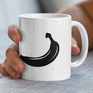 Könnte beinhalten: Weiße Keramik-Tasse mit einer schwarzen Bananensilhouette. Die Tasse hat einen gebogenen Henkel und eine glänzende Oberfläche. Das Bananen-Design ist eine einfache, kräftige Grafik. Die Tasse wird von einer Person gehalten.