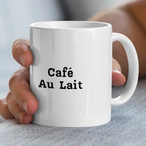 May include: White ceramic coffee mug with the words "Café Au Lait" printed in black. The mug has a curved handle and is held by a person.