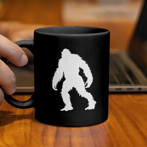 Könnte beinhalten: Schwarze Keramik-Tasse mit einer weißen Bigfoot-Silhouette. Die Tasse hat einen gebogenen Griff und eine glänzende Oberfläche. Das Bigfoot-Design ist mittig auf der Tassenoberfläche platziert und erzeugt einen starken Kontrast. Die Tasse wird von einer Person gehalten.
