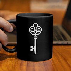 Könnte beinhalten: Schwarze Keramik-Tasse mit einem weißen, verzierten Schlüsseldesign. Der Schlüssel hat eine detaillierte, antike Ästhetik. Die Tasse wird von einer Person gehalten, im Hintergrund ist ein Laptop zu sehen. Die Tasse steht auf einer Holzoberfläche.