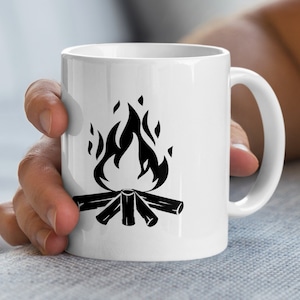 Könnte beinhalten: Weiße Keramiktasse mit einer schwarzen Lagerfeuergrafik. Das Design zeigt Flammen über einem Holzstapel. Die Tasse hat einen gebogenen Henkel und wird von einer Person gehalten.