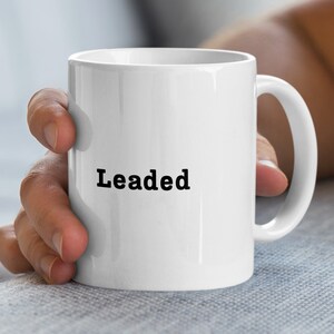 Peut inclure: Une tasse en céramique blanche avec le mot "Leaded" imprimé en noir. La tasse a une anse incurvée et est tenue par une personne. La tasse est conçue pour les boissons chaudes ou froides.