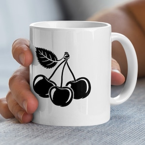 Könnte beinhalten: Weiße Keramiktasse mit einem schwarzen Kirschdesign. Das Design zeigt drei Kirschen und ein Blatt. Die Tasse hat einen gebogenen Henkel und wird von einer Hand gehalten.
