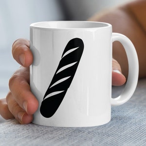 Könnte beinhalten: Weiße Keramik-Tasse mit einer schwarzen Abbildung eines Baguettes. Die Tasse hat einen gebogenen Henkel. Das Design ist schlicht und modern, geeignet für Kaffee oder Tee.