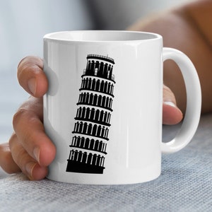 Könnte beinhalten: Weiße Keramiktasse mit einer schwarzen Silhouette des Schiefen Turms von Pisa. Die Tasse hat einen gebogenen Henkel. Das Design ist eine einfache, kräftige Grafik. Die Tasse wird von einer Person gehalten.