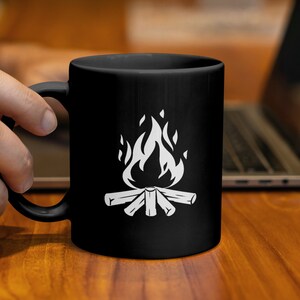 Könnte beinhalten: Schwarze Keramik-Tasse mit weißer Lagerfeuer-Grafik. Das Design zeigt Flammen über Holzscheiten. Die Tasse wird von einer Hand gehalten. Im Hintergrund ist ein Laptop zu sehen.
