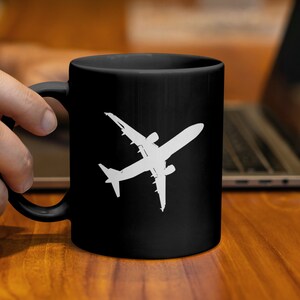 Könnte beinhalten: Schwarze Keramik-Tasse mit einer weißen Flugzeug-Silhouette. Die Tasse wird von einer Hand gehalten, mit einem Laptop im Hintergrund. Die Tasse steht auf einer Holzoberfläche.