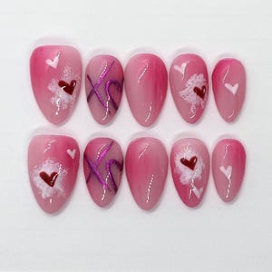 May include: Set of ten pink and white false nails with different Valentine's Day themed designs. Some nails have pink and white hearts, others have pink and white swirls, and others have a pink and white X.