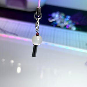 May include: A small charm featuring a white, spherical bead and a black, cylindrical base. It's connected to a silver clasp and a pink cord. The background is blurred, with purple and blue hues. A decorative item.