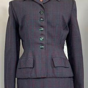 Könnte beinhalten: Ein Vintage-Anzug mit grauem Karomuster und passender Jacke und Rock. Die Jacke hat eine taillierte Passform, gekerbte Revers und dekorative grüne Knöpfe. Der Rock ist im klassischen Bleistiftstil gehalten. Der Anzug ist aus einem gewebten Stoff gefertigt.