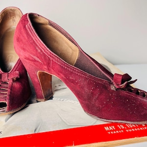 Puede incluir: Un par de zapatos de tacón peep-toe burdeos vintage. Los zapatos presentan un detalle de lazo en la pala y un tacón de altura media. Los zapatos están hechos de un material similar al ante y se exhiben en una revista vintage.