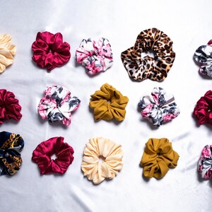 Pode incluir: Conjunto de 14 scrunchies para cabelo de cores variadas e padrões diferentes, incluindo flores, estampa de leopardo e cores sólidas. Os scrunchies estão dispostos sobre um fundo branco.