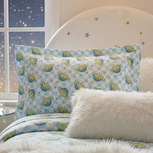 May include: A light blue pillowcase with a repeating pattern of green and yellow cartoon aliens on a checkered background. The pillowcase is part of a bedding set, with a fluffy white pillow in the foreground.
