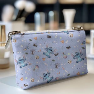 May include: A light blue cosmetic bag with a zip closure. The bag features a repeating pattern of a blue cartoon character, ghosts, trees, and the word "Boo". The bag is rectangular and sits on a white surface.
