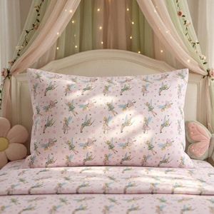 Puede incluir: Juego de funda de almohada y sábanas rosas con un patrón repetido de hadas y estrellas. Las hadas visten trajes verdes y rosas. La ropa de cama está sobre una cama con dosel y luces de hadas.