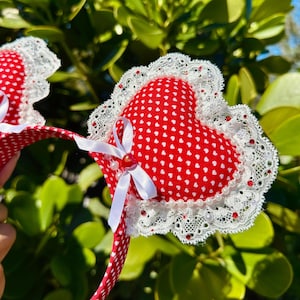 Peut inclure: Un serre-tête rouge orné de cœurs. Chaque cœur est rouge à pois blancs, bordé de dentelle blanche et de petites perles rouges. Des nœuds blancs et des perles ornent le centre des cœurs. Le serre-tête est rouge à pois blancs.