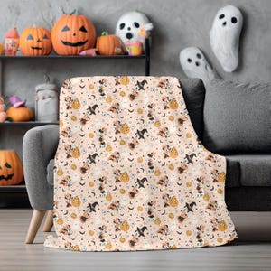 Op de afbeelding: Een Halloween-deken met een herhalend patroon van stripfiguren met heksenhoeden, pompoenen, vleermuizen en grafstenen op een lichtperzik achtergrond. De deken is gedrapeerd over een grijze bank, met Halloween-decoraties op de achtergrond.