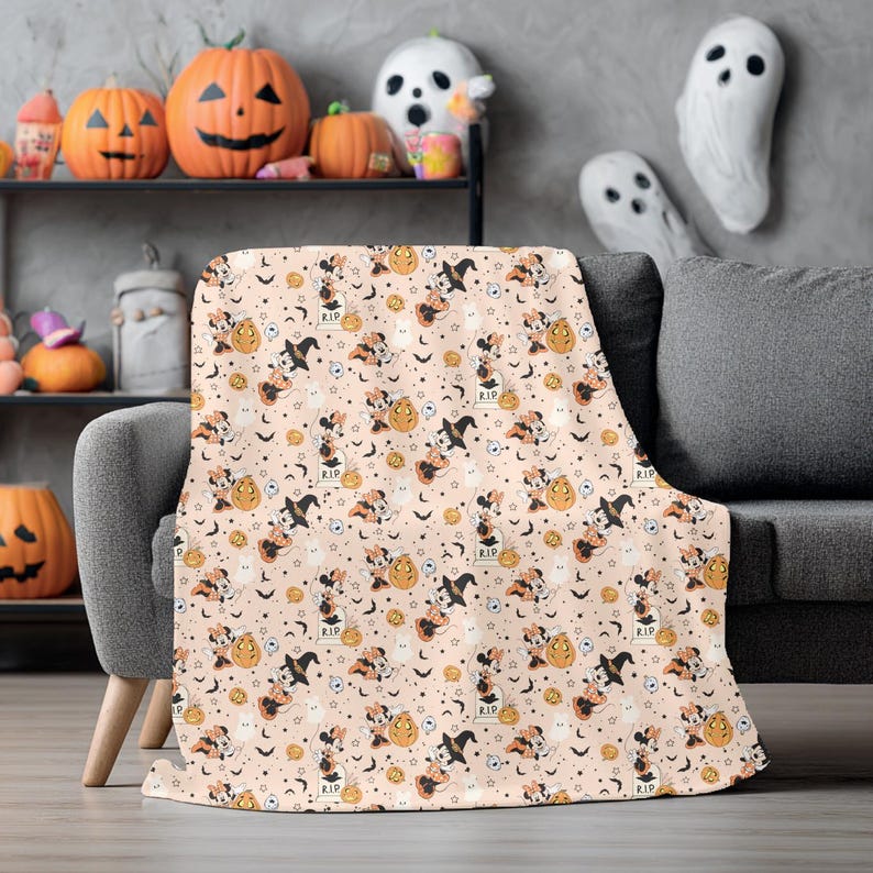 Op de afbeelding: Een Halloween-deken met een herhalend patroon van stripfiguren met heksenhoeden, pompoenen, vleermuizen en grafstenen op een lichtperzik achtergrond. De deken is gedrapeerd over een grijze bank, met Halloween-decoraties op de achtergrond.