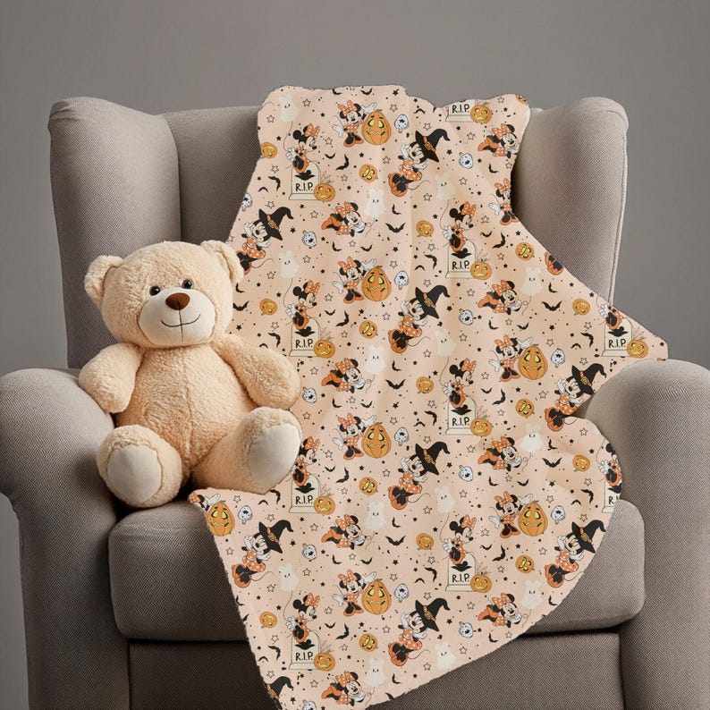 Op de afbeelding: Een beige teddybeer zit op een grijze fauteuil naast een Halloween-deken. De deken heeft een herhalend patroon van Minnie Mouse met een heksenhoed, pompoenen, vleermuizen en grafstenen op een lichtperzikkleurige achtergrond.