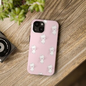 Könnte beinhalten: Eine rosa Handyhülle mit einem sich wiederholenden Muster aus weißen Katzenillustrationen mit Schleifen. Die Hülle hat einen schwarzen Kameraobjektiv-Ausschnitt und liegt auf einer Holzoberfläche. Eine Kamera und eine Pflanze sind im Hintergrund zu sehen.