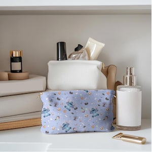 May include: A light blue cosmetic bag with a gold zip, featuring a repeating pattern of a cartoon character, trees, and the word "Boo". The bag is on a white shelf with other cosmetic items.