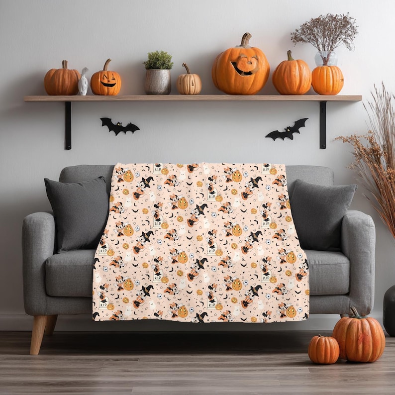 Op de afbeelding: Een Halloween-thema deken over een grijze bank. De deken heeft een herhalend patroon van pompoenen, geesten, vleermuizen en heksenfiguren op een lichtroze achtergrond. Oranje pompoenen en decoraties staan op een plank boven de bank.