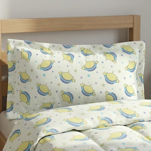 May include: A light-colored pillowcase and comforter set featuring a repeating pattern of yellow alien characters with blue and purple accents, and small stars. The bedding is on a wooden bed frame.