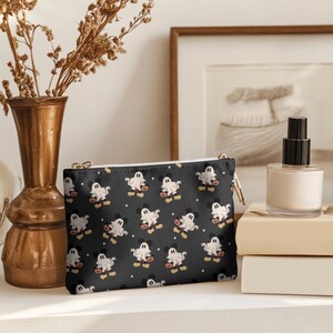 May include: Black zippered pouch with a repeating pattern of cartoon ghosts holding jack-o'-lanterns. The ghosts have Mickey Mouse ears and are set against a black background with small white stars. A gold zipper and pull complete the design.