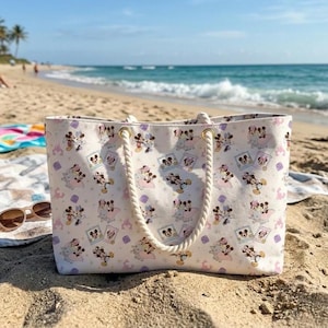 May include: A white tote bag with a repeating pattern of cartoon characters in pink, purple, and yellow. The bag has rope handles and gold-colored eyelets. The bag is sitting on a sandy beach with sunglasses.