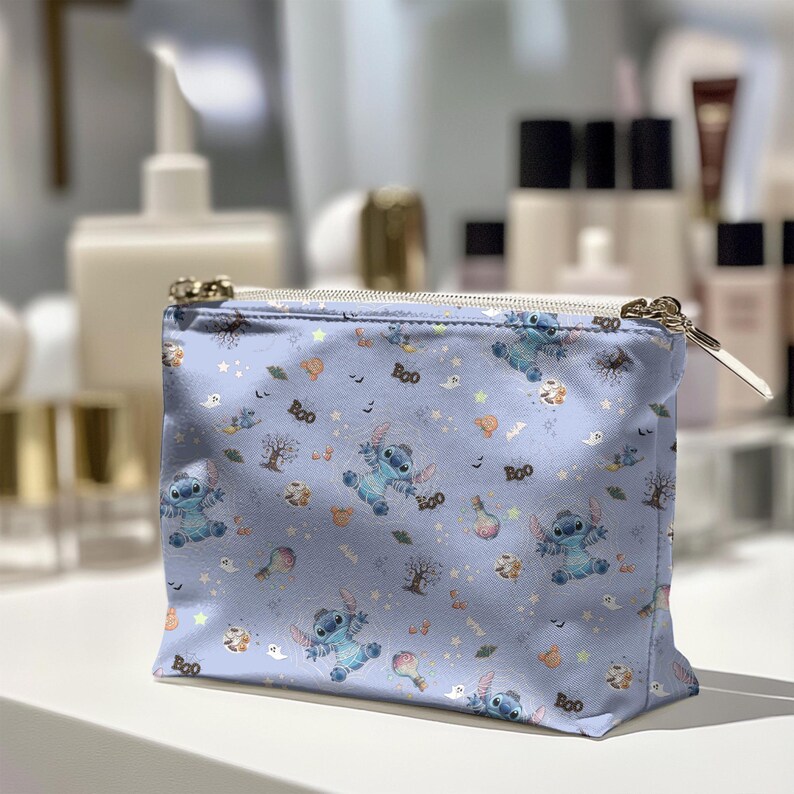 May include: A pale blue cosmetic bag featuring a pattern of Stitch, ghosts, trees, and the word "Boo". The bag has a gold zip and a white zip pull. The bag is placed on a white surface.