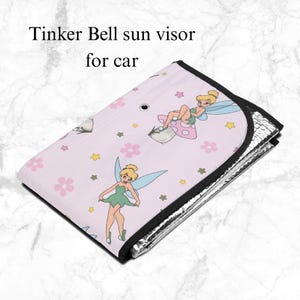 May include: A folded car sun visor featuring a light pink background with a repeating pattern of Tinker Bell, pink flowers, and yellow stars. The text Tinker Bell sun visor for car is printed on the surface. It has a black border and silver lining.