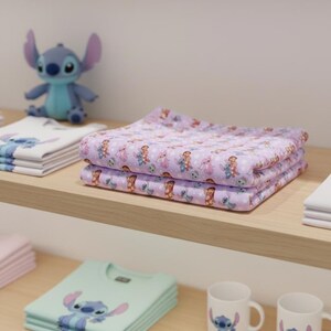 Peut inclure: Tissu lavande pli&eacute; avec un motif r&eacute;p&eacute;titif de personnage de dessin anim&eacute;. Une figurine bleue de personnage de dessin anim&eacute; est sur l'&eacute;tag&egrave;re derri&egrave;re le tissu. D'autres articles sur l'&eacute;tag&egrave;re comprennent des t-shirts pli&eacute;s et des tasses.