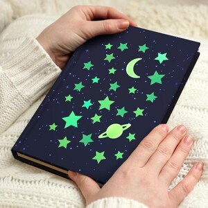 May include: A dark blue journal with a celestial design featuring green stars, a crescent moon, and a planet. The journal is held by a person wearing a white sweater and blue jeans. The journal is resting on a white pillow.