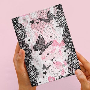 May include: A notebook with a pink background and a design featuring butterflies, hearts, and lace. The notebook is framed with a black and white checkered lace pattern. The butterflies are black with detailed wing patterns.