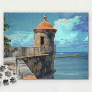 Puede incluir: Una torre de piedra con un techo naranja se alza sobre un muro de piedra con vistas a una masa de agua azul. La torre tiene una pequeña ventana y una barandilla. El cielo es azul con nubes blancas.