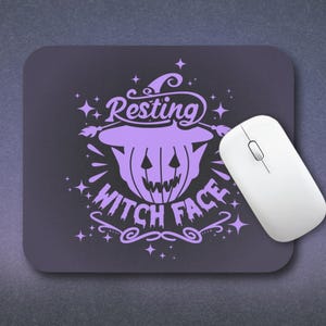 Op de afbeelding: Een paarse muismat met een Halloween-thema. Het ontwerp bevat een pompoen met de tekst "Resting Witch Face" in een decoratief lettertype, samen met sterren en krullen. Een witte draadloze muis ligt naast de muismat.