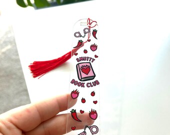 Peut inclure: Marque-page transparent en acrylique avec un pompon rouge. Le marque-page présente des illustrations de piments, de cœurs, de menottes et d'un livre avec un cœur. Le texte "Smutty Book Club" est imprimé sur le marque-page.