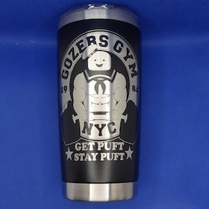 May include: A black stainless steel tumbler with a white logo that says "Gozers Gym 1984 NYC Get Puft Stay Puft". The logo features a cartoon character with a sailor hat and a muscular physique.